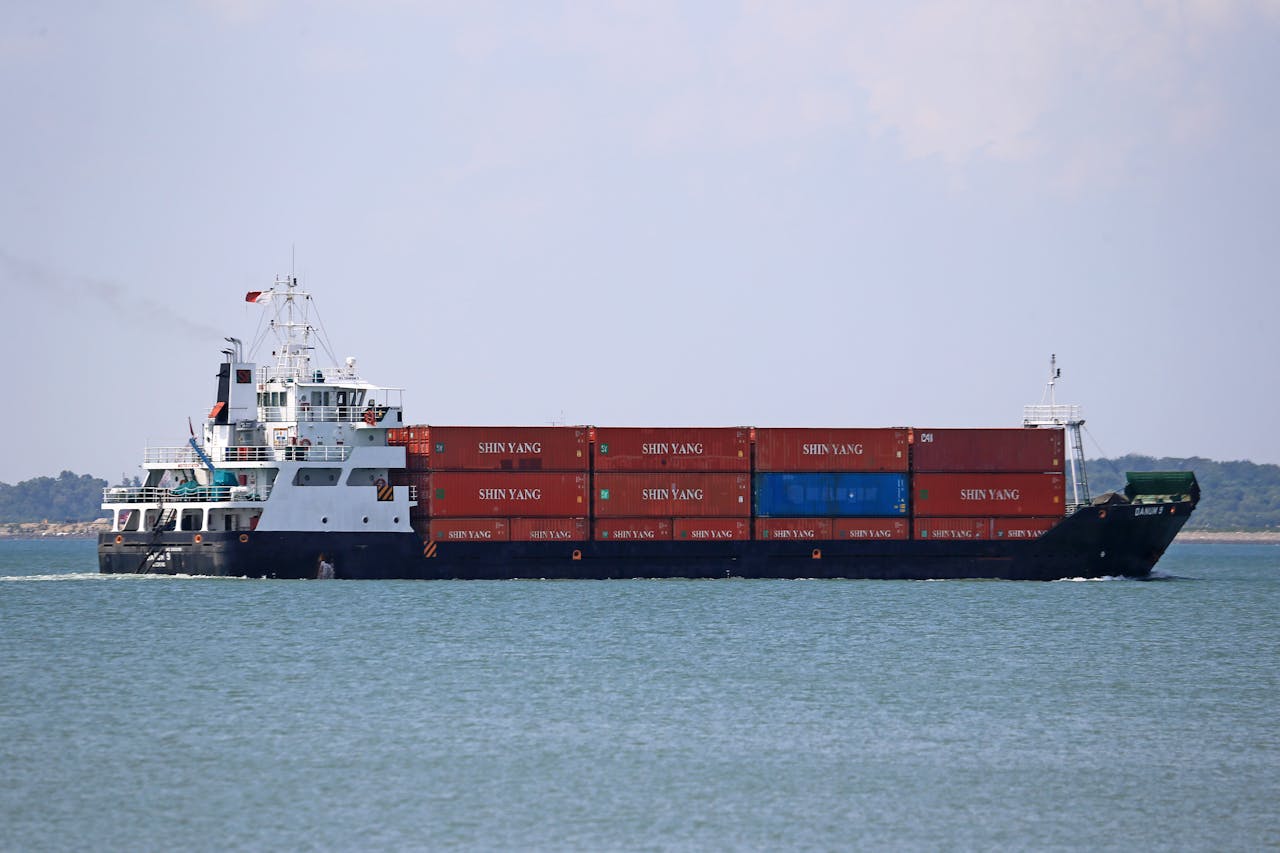 A cargo ship carrying shipping containers sails across the ocean under bright daylight.