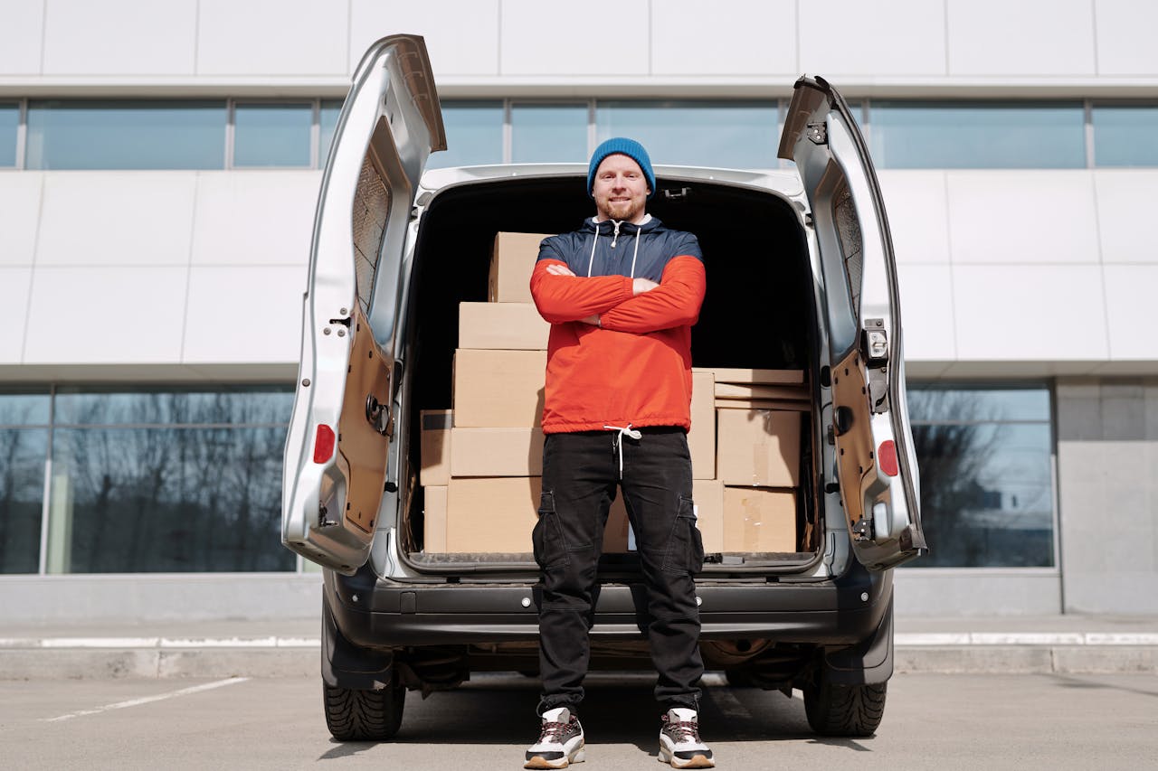 Man in red jacket and blue beanie stands by van of boxes in the city, ready for delivery.